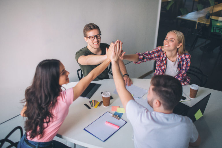 Happy and cheerful young people sit around table and keep hands together. They look happy and joyful. People play as team in room